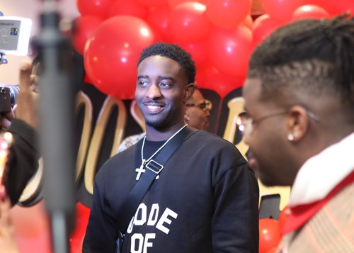visual storytelling for brand transformation. Man in a black “Code of” sweatshirt smiling during a red carpet media event, surrounded by cameras and vibrant red balloons.